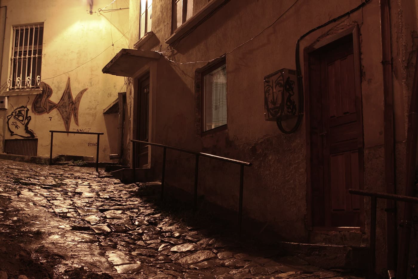 Nighttime street scene with lamplight illuminating pavement, building facades, and visible graffiti on walls