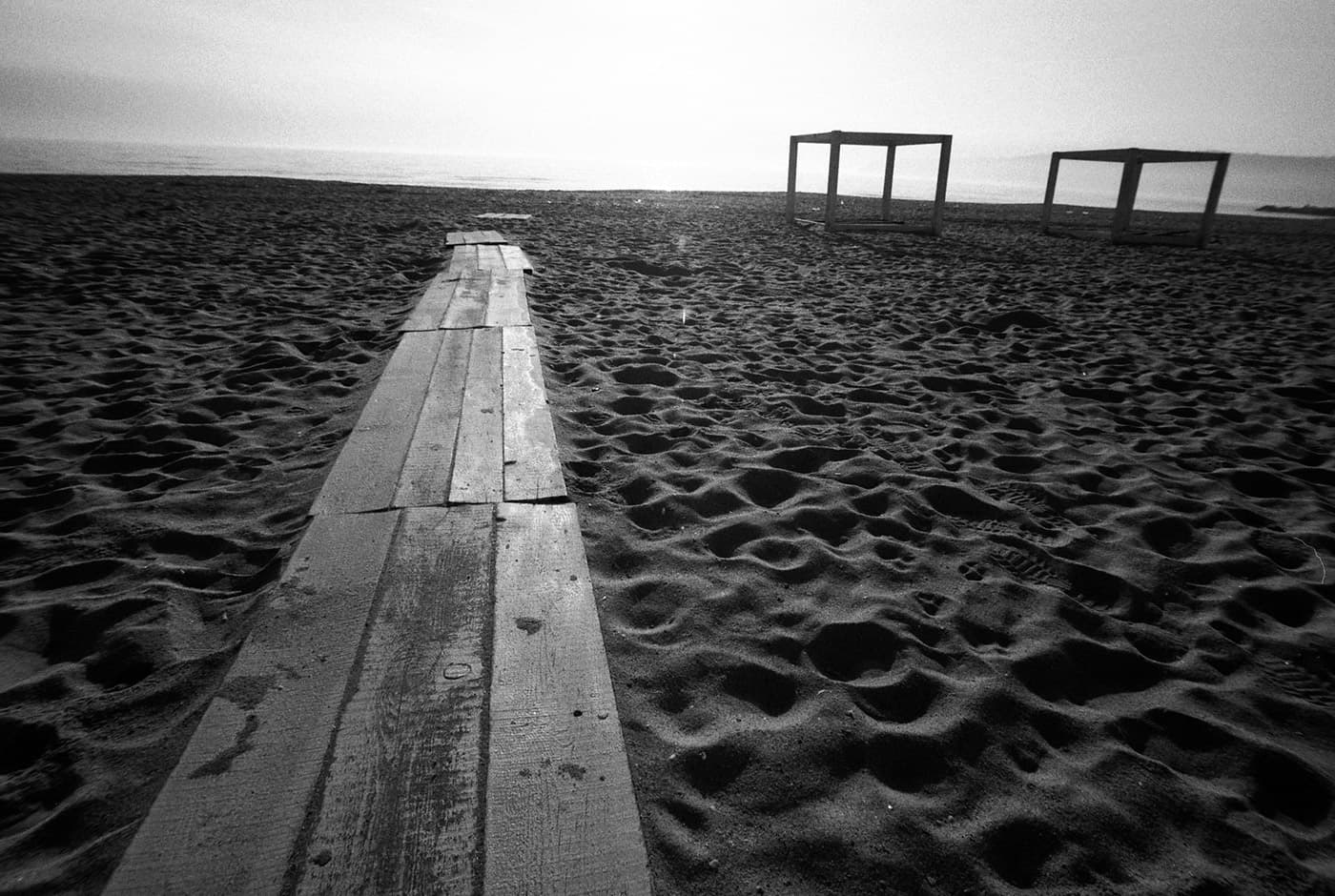 An empty beach with a wooden pathway leading toward the sea and a couple of wooden cube structures nearby.