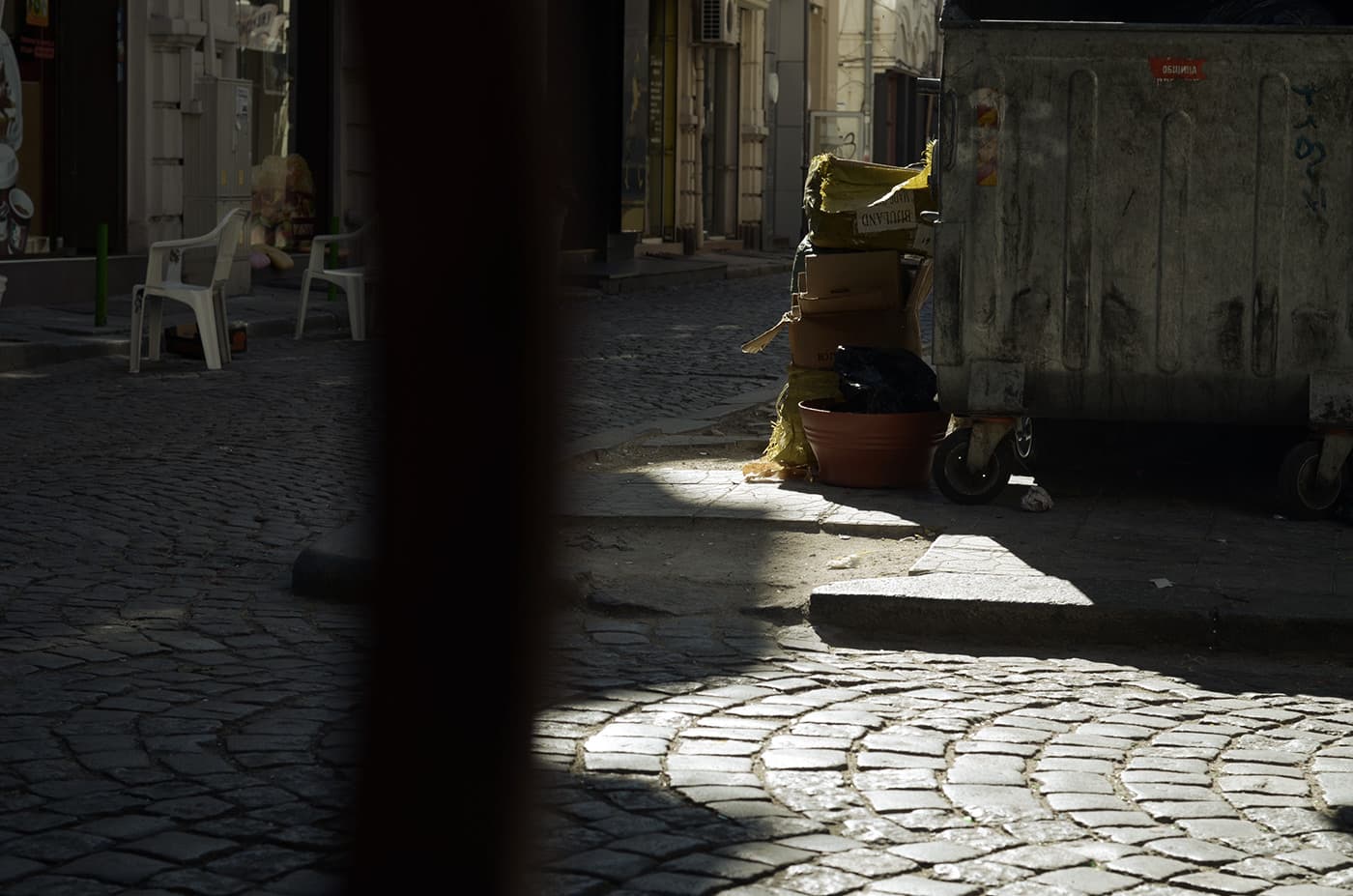 Street scene with pavement, a trash can surrounded by various objects, boxes, and chairs placed along the sidewalk