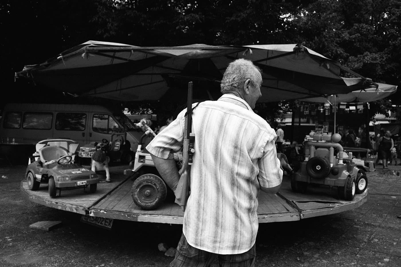 A man standing in front of a children’s carousel holding a plastic shotgun on his shoulder