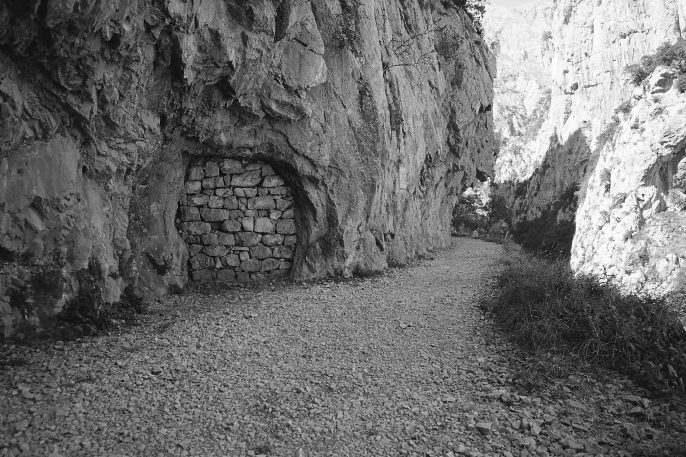A rocky mountain path with a cave or portal-like opening in the rocks that is blocked by a rock wall