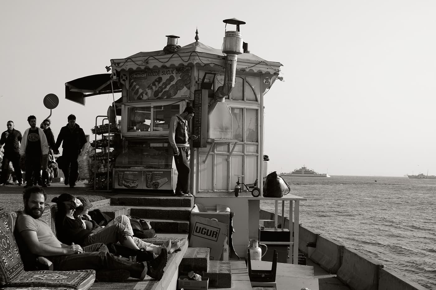 Food pavilion next to the sea with people sitting on carpets and cushions arranged on concrete steps along the waterfront