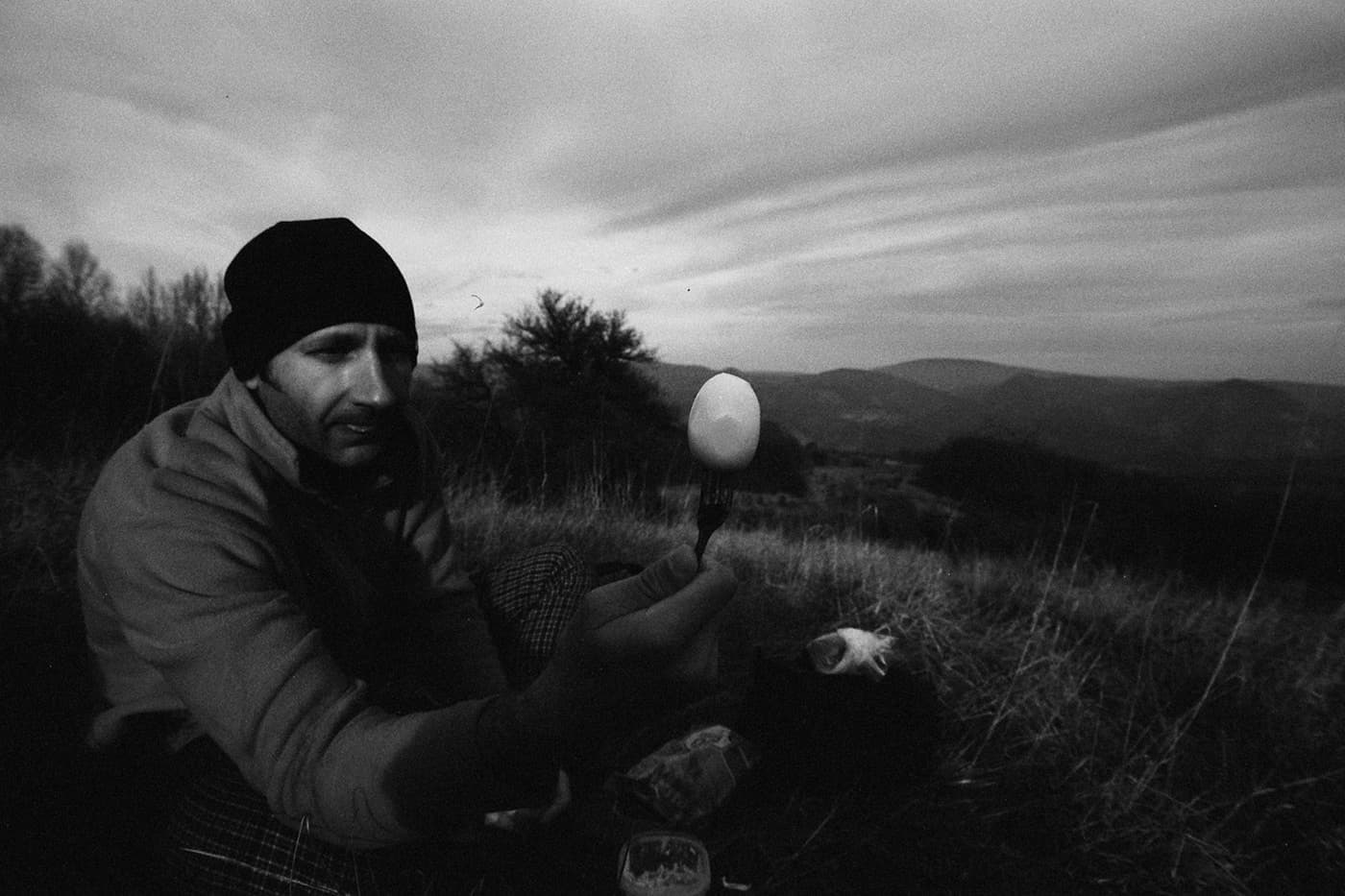 A man sitting on the ground in a mountain field holding a fork with a boiled egg, with mountains in the background.