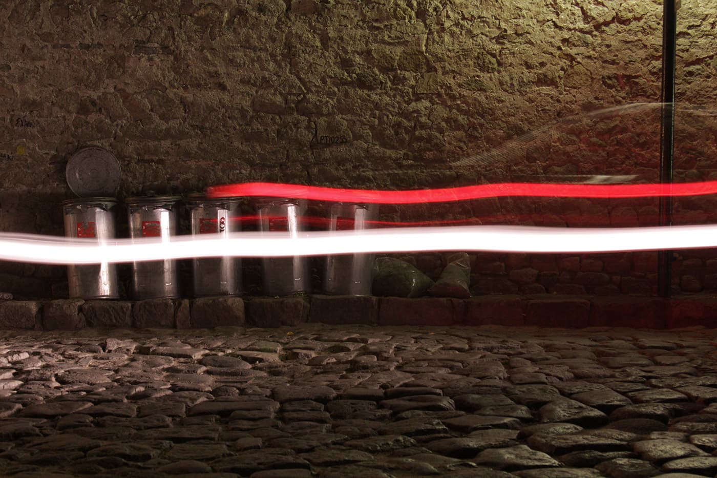 Nighttime long exposure showing light trails from a passing car in front of trash cans arranged in a row along the street