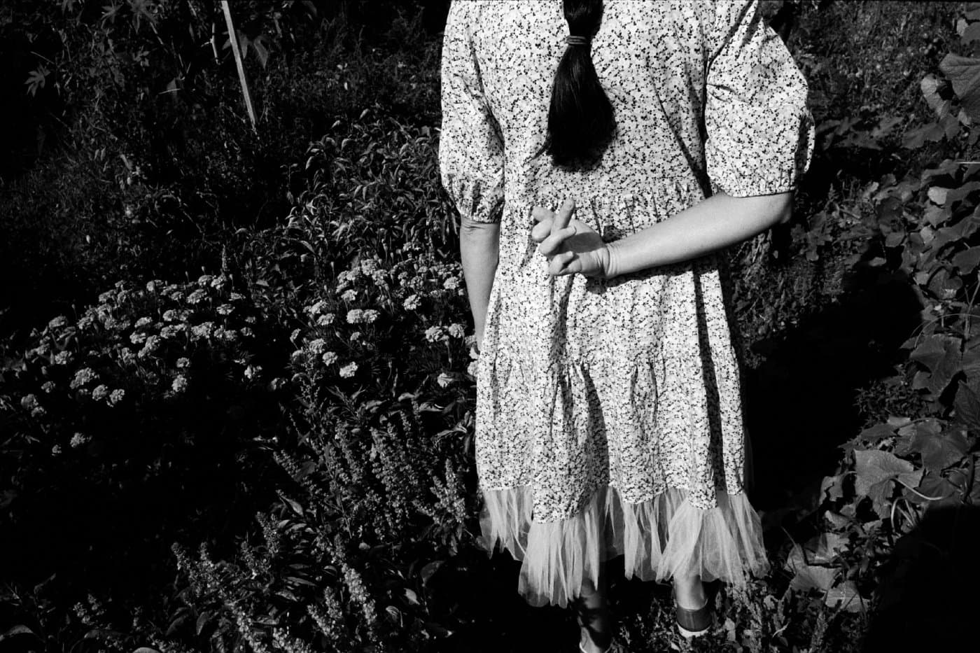 A girl standing in a garden with her back to the camera, holding her hand behind her showing a three-finger salute toward the photographer