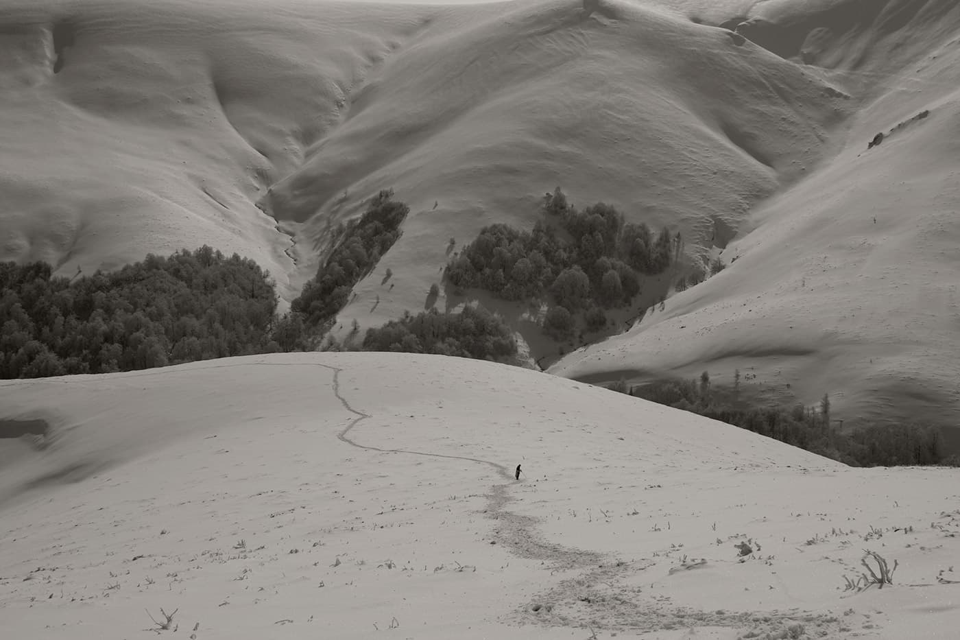 Mountain landscape with a snow-covered path leading into the distance where a single person is walking, with snowy hills, slopes, and trees in lower areas