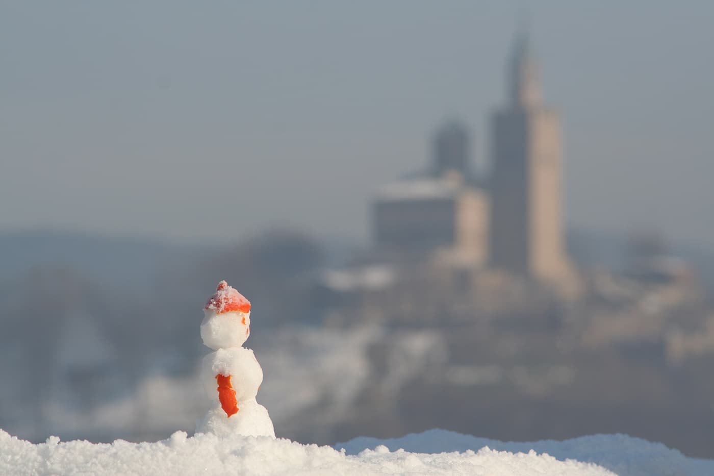 Small snowman made of snow with orange peels used for hat, hands, and face, with Veliko Tarnovo Tsarevets fortress blurred in the background