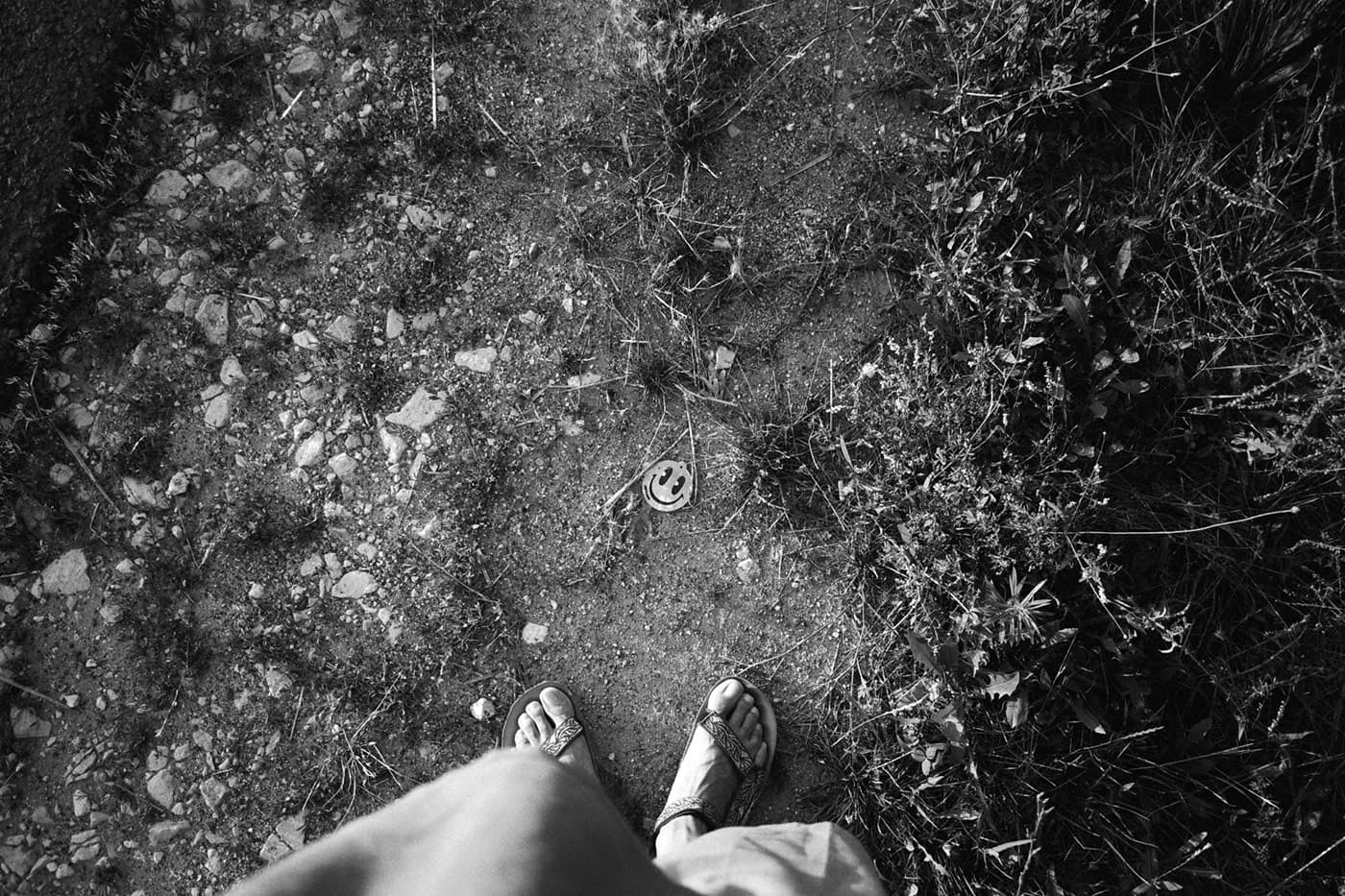 A man's feet next to a smile emoji sticker lying on a rural path.
