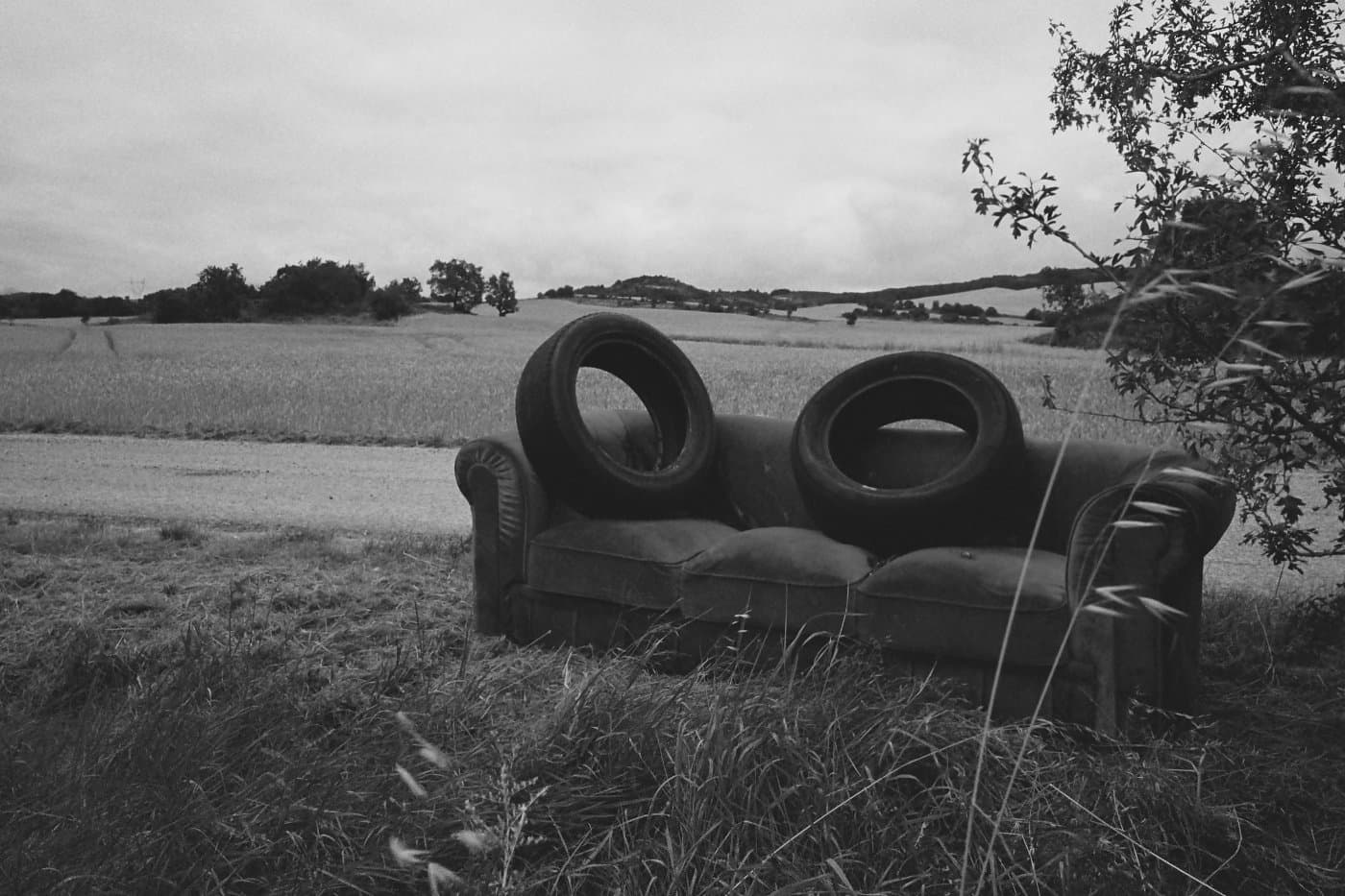 Abandoned sofa in an outdoor field beside wheat crops with two automobile tires resting on top