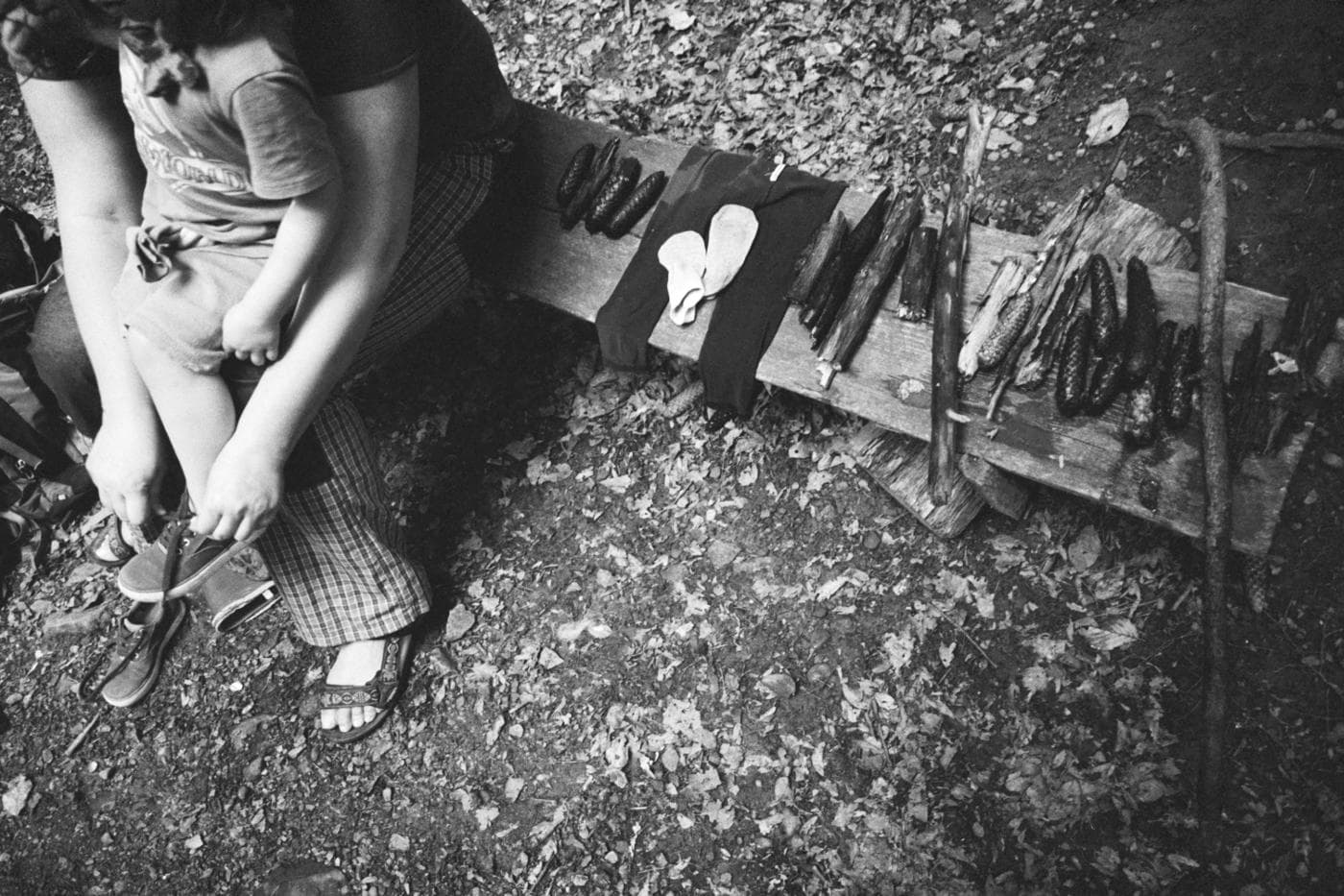 A woman holding a small child and putting on their shoes on a bench with neatly arranged forest objects like sticks, pine cones, socks, and cloth