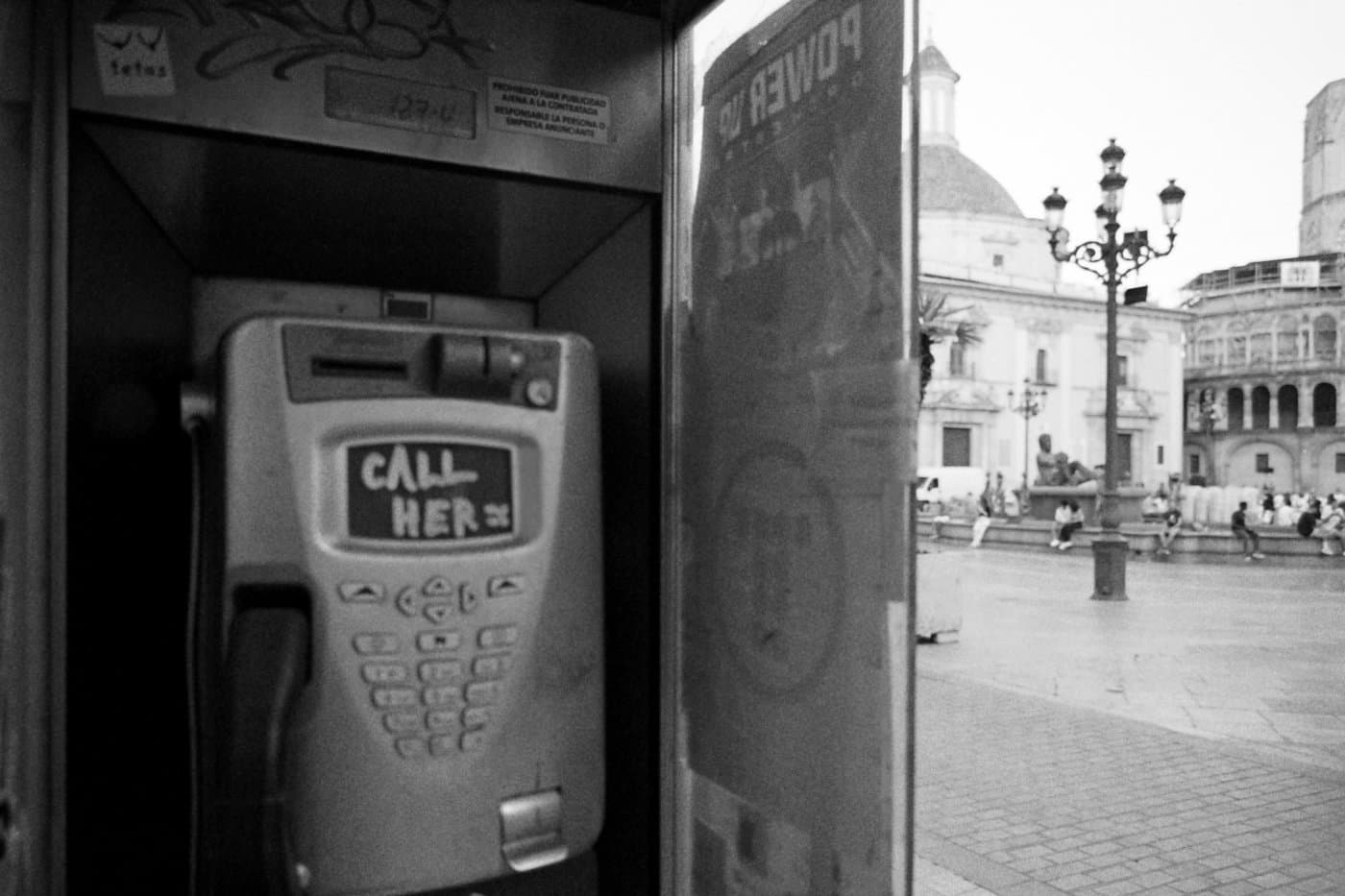 A street phone booth showing the phone inside and part of a city square, with the words call her and tetas written on the phone