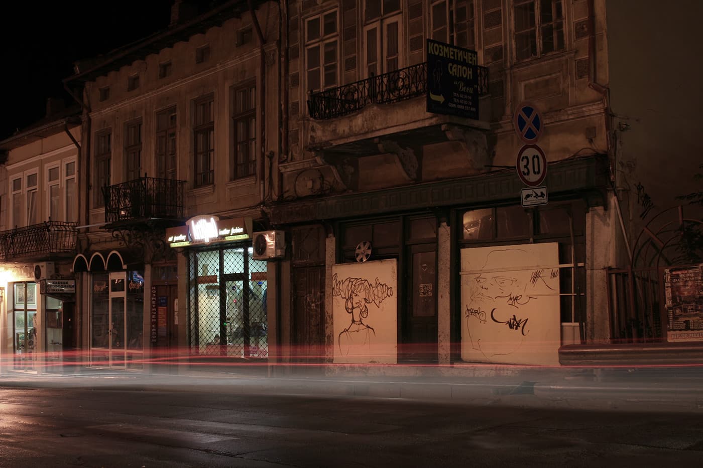 Nighttime long exposure of a city street with car light trails, buildings along the road, and street stencils on some building facades
