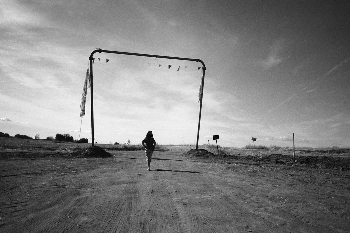 A distant figure walking through a dry field near a simple portal made of poles with signs placed in the ground nearby