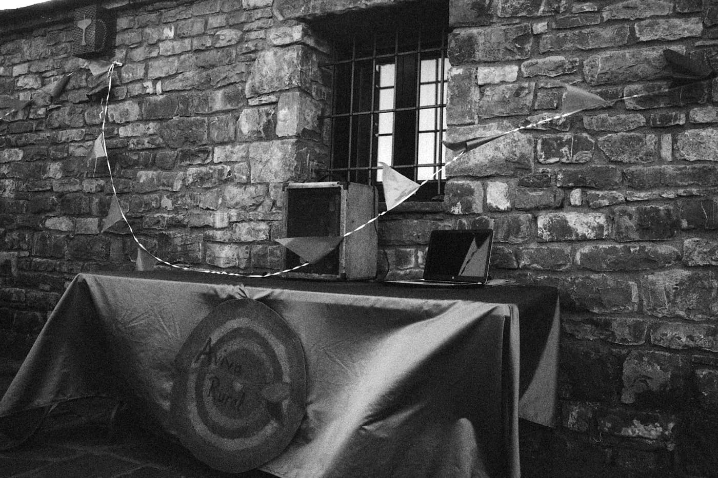 Black and white photo of a table covered with a shiny fabric next to a stone wall, with a sign reading "aviva rural", a laptop, a wooden box, and triangular flags on a string above a window