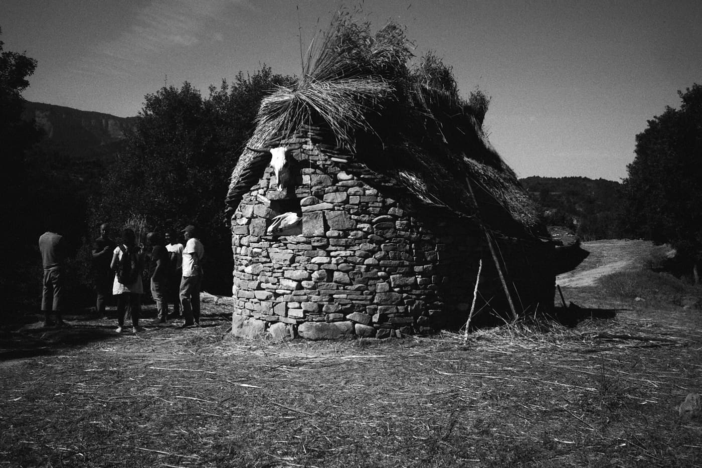 Group of people standing next to a round stone house with a straw roof and a cow skull mounted above a window